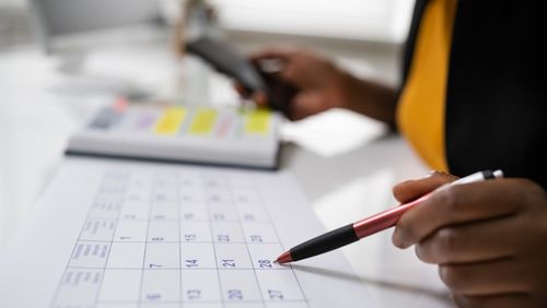 Eine Frau sitzt vor ener Tastatur und einem Kalender, zu sehen ist im Vordergrund eine Hand mit Stift und im Hintergrund eine Hand mit einem Handy.